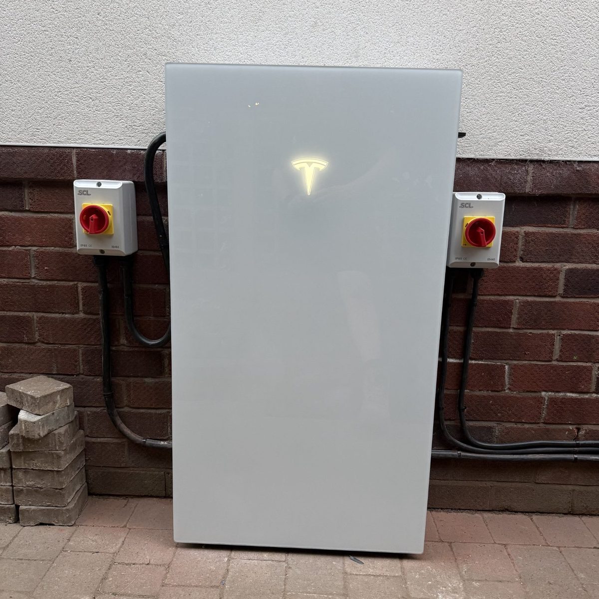 A Tesla Powerwall in place against a brick house wall, with an isolator switch and conduit either side